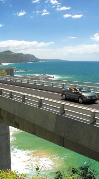 Long view of car driving along Sea Cliff Bridge, Grand Pacific Drive, with coastal views in background, Illawarra
