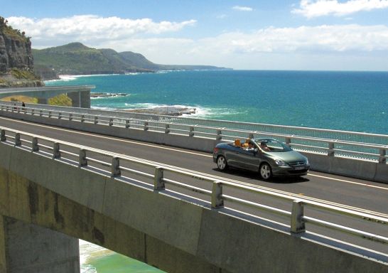 Long view of car driving along Sea Cliff Bridge, Grand Pacific Drive, with coastal views in background, Illawarra